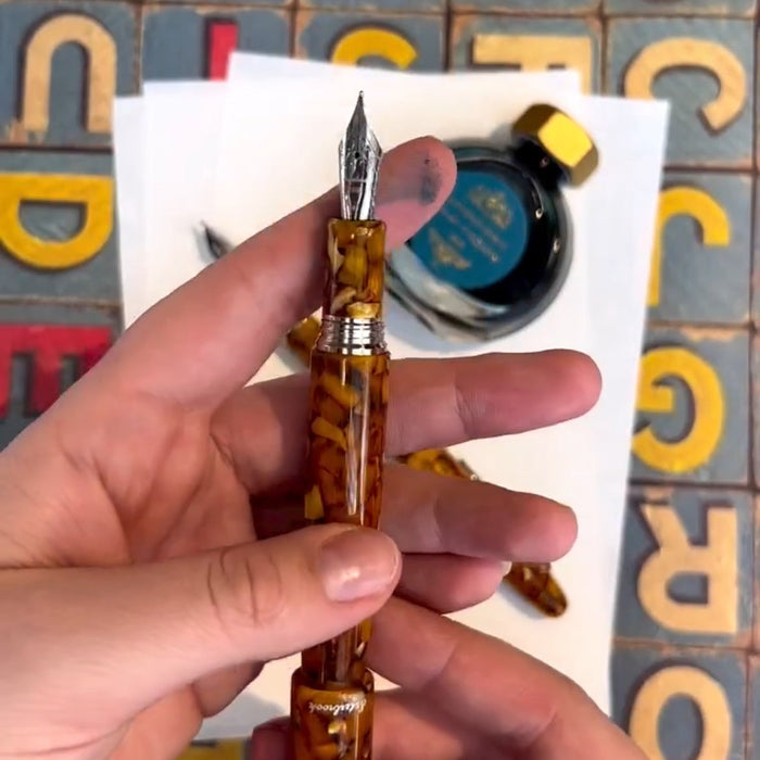 hand holding a fountain pen with decorative brown body with silver nib along with a bottle of ink, white paper, and alphabet letters in the background