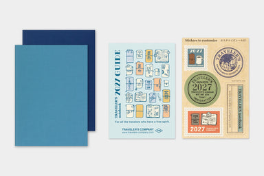 TRAVELER'S notebook Refill- Passport Size- 2027 Weekly Diary displayed on a light gray background, featuring two blue Diaries, a light blue card labeled 'Travelers' 2027 Guide', and one sticker sheet with travel-themed labels.