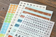 Three sheets of planner stickers laid out on a rustic wooden table. The sheets include colorful round stickers in orange, yellow, green, and blue, black-and-white alphabet and number stickers, and icon stickers for travel and calendar purposes.