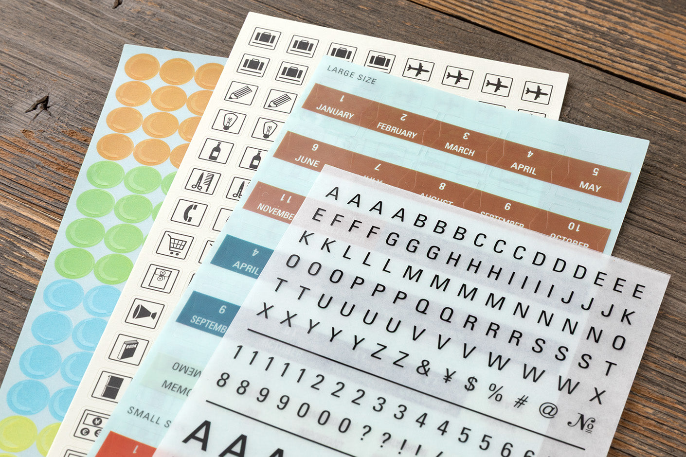 Three sheets of planner stickers laid out on a rustic wooden table. The sheets include colorful round stickers in orange, yellow, green, and blue, black-and-white alphabet and number stickers, and icon stickers for travel and calendar purposes.