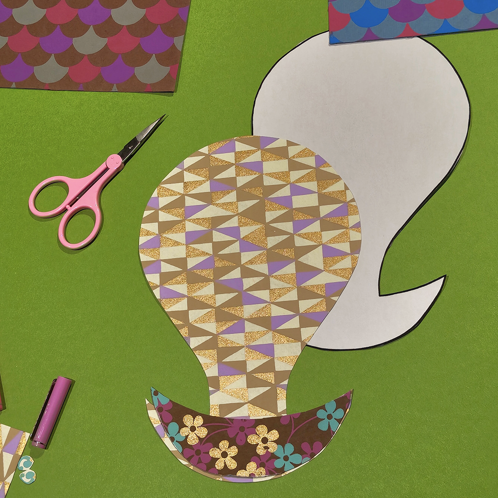 Colorful paper cutout of a hot air balloon with geometric patterns on a green background, accompanied by scissors.