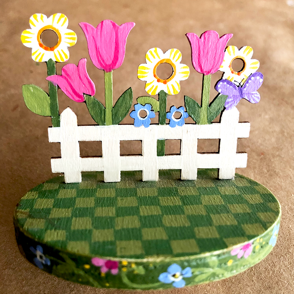 Decorative wooden garden scene with flowers and a white picket fence on a checkered base.