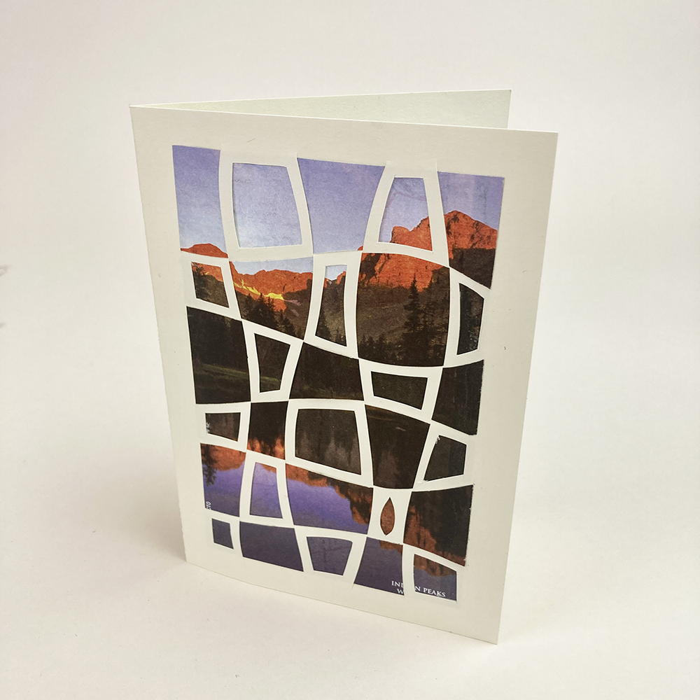 Greeting card with mountain and lake design on a white background with cut paper geometric pattern laid on top.