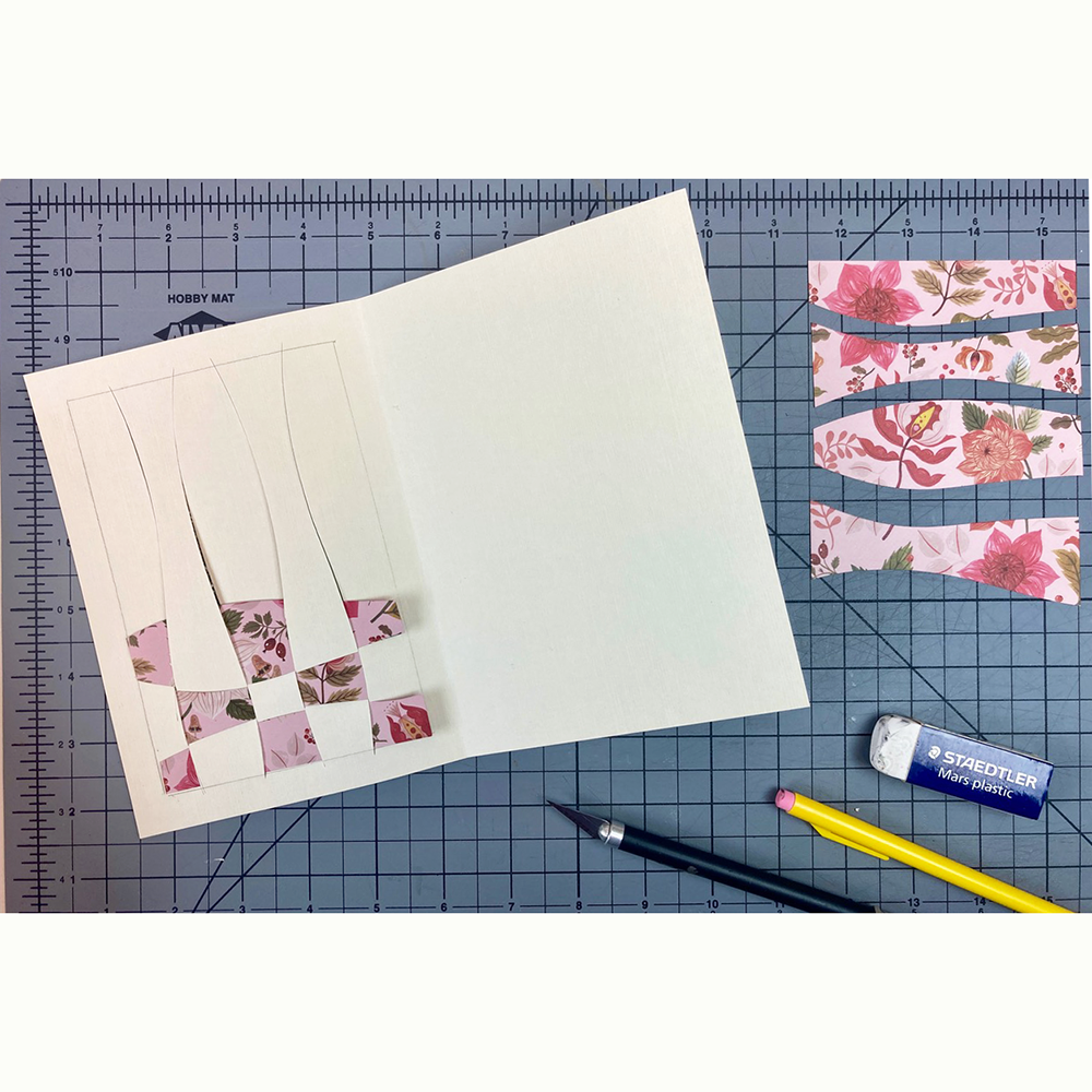 Woven paper design on a white card in progress on a cutting mat background with art supplies