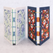 Two decorative hardcover books standing upright side by side on a light marble surface against a white wall. The left book has a cream cover with blue floral linocut-style illustrations and blue thread binding.