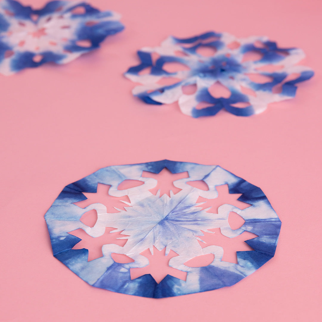 3 paper snoflakes dip-dyed in indigo ink displayed on a pink background