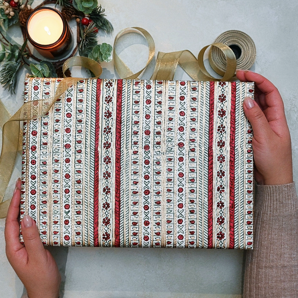 Gift box with decorative ribbon held by hands, with a candle and greenery in the background