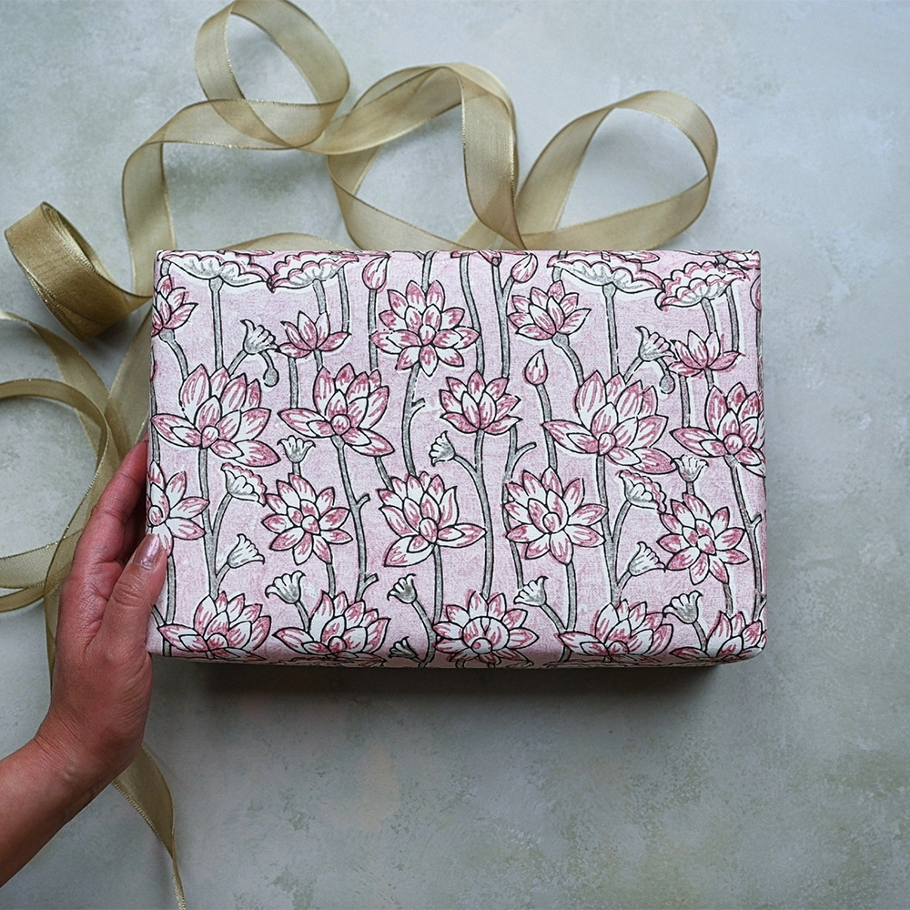 Handmade Indian Cotton Paper- Block Printed lotus floral pattern with pink flowers and green stems used as gift wrap. Shown with a hand and green ribbon.