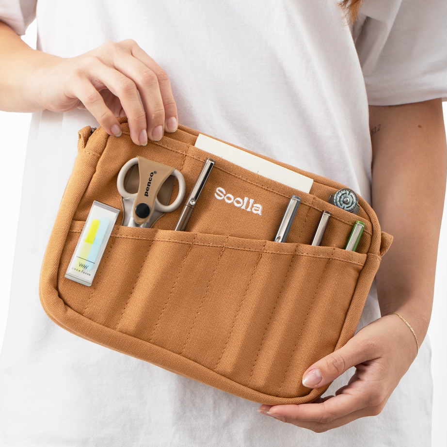 A person in a white shirt holds an open tan canvas organizer pouch filled with pens, scissors, and small accessories. The pouch has multiple pockets and the brand name 'Soolla' printed on the front.