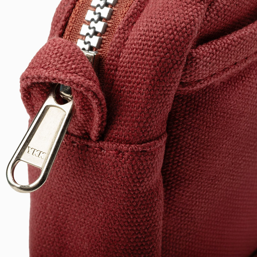 Close-up of a burgundy canvas bag featuring a silver metal zipper pull and a matching fabric loop tab, with a white background visible in the corner.