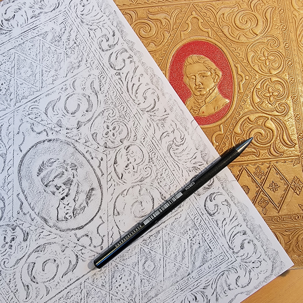 White paper graphite rubbing  with pencil sitting on decorative embossed book cover, featuring intricate patterns and portrait.