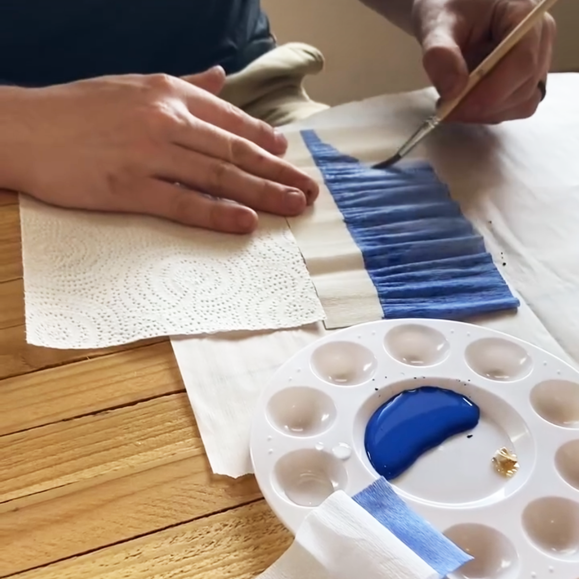 Person painting a blue stripe on a white crepe paper with a brush, next to a paint palette.