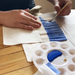 Person painting a blue stripe on a white crepe paper with a brush, next to a paint palette.