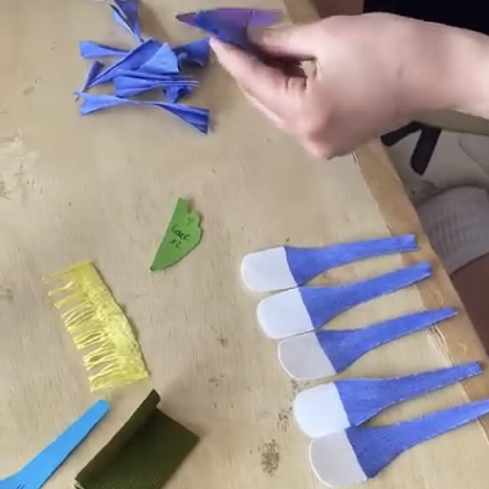 Hand arranging blue paper petals on a wooden surface with other craft materials.