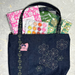 A dark indigo denim tote bag with white embroidered floral patterns and a pink patch,  shown filled with various colorful stationery and craft items on a textured gray background.